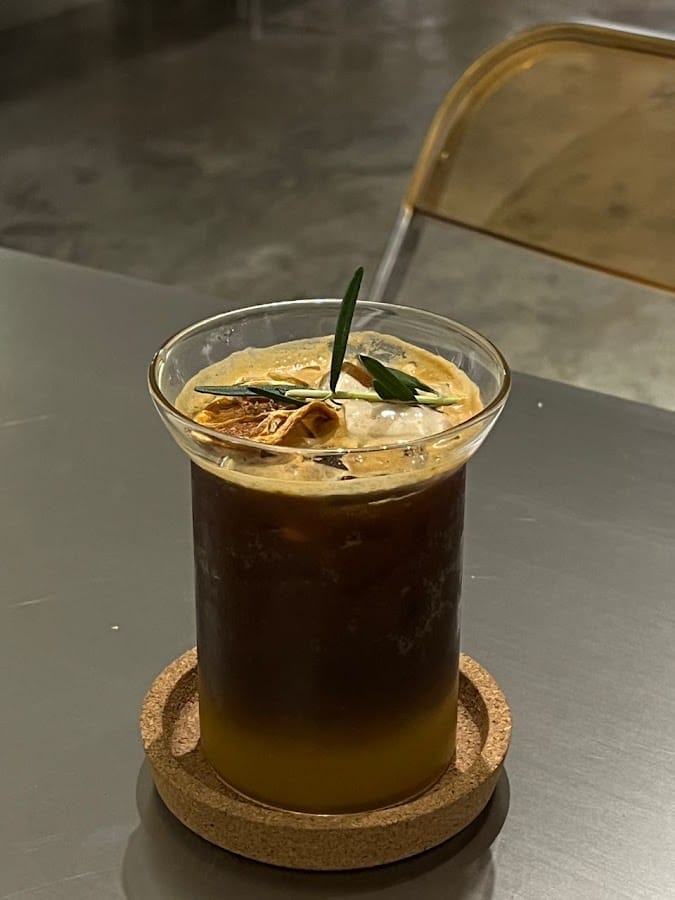 An iced coffee layered over orange, with a dried orange slice and a sprig of herbs